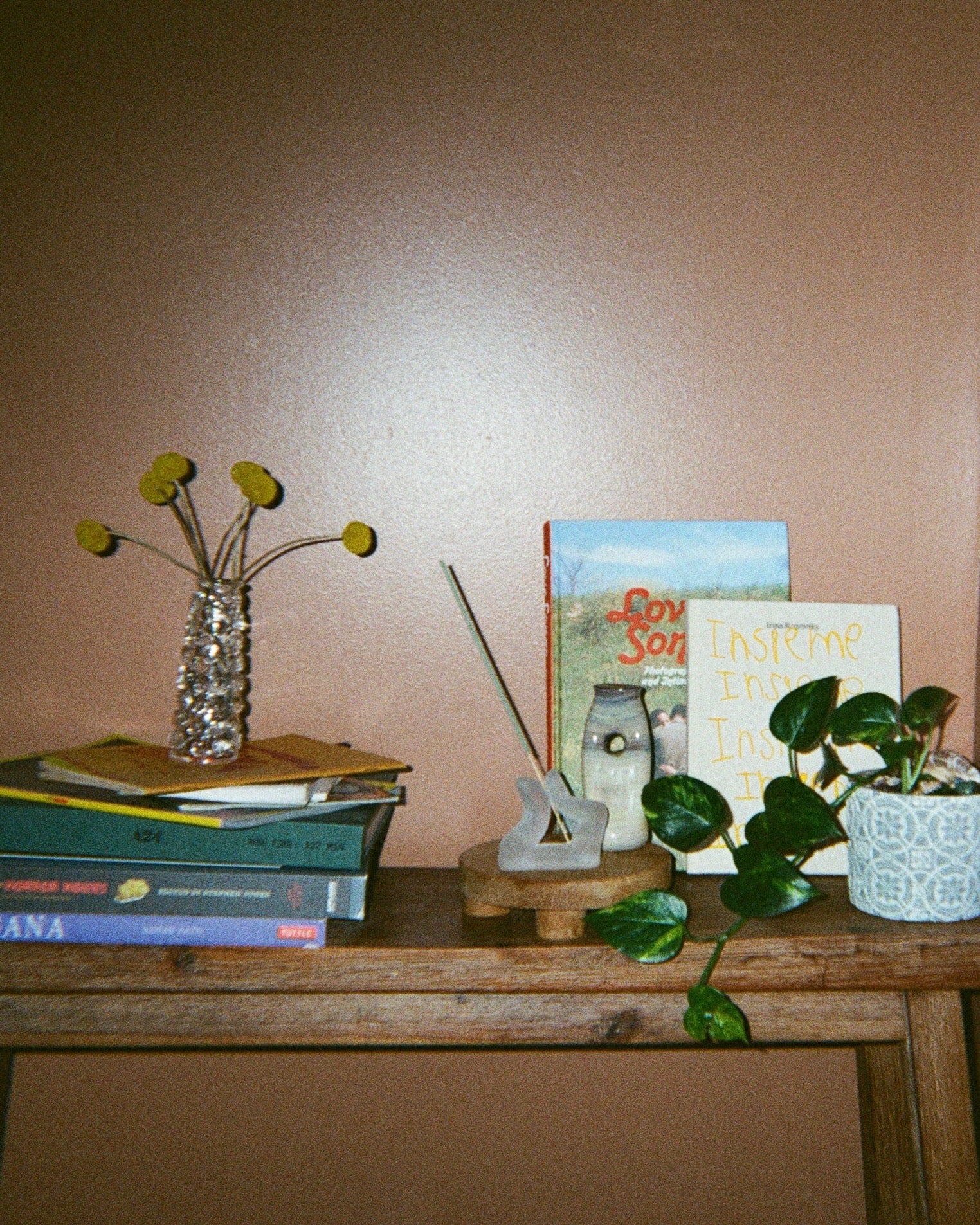 A clear glass flower vessel holding yellow billy buttons, a finger incense holder, another small vessel, a plant, and stacked books on a wooden bench against a brown wall. 