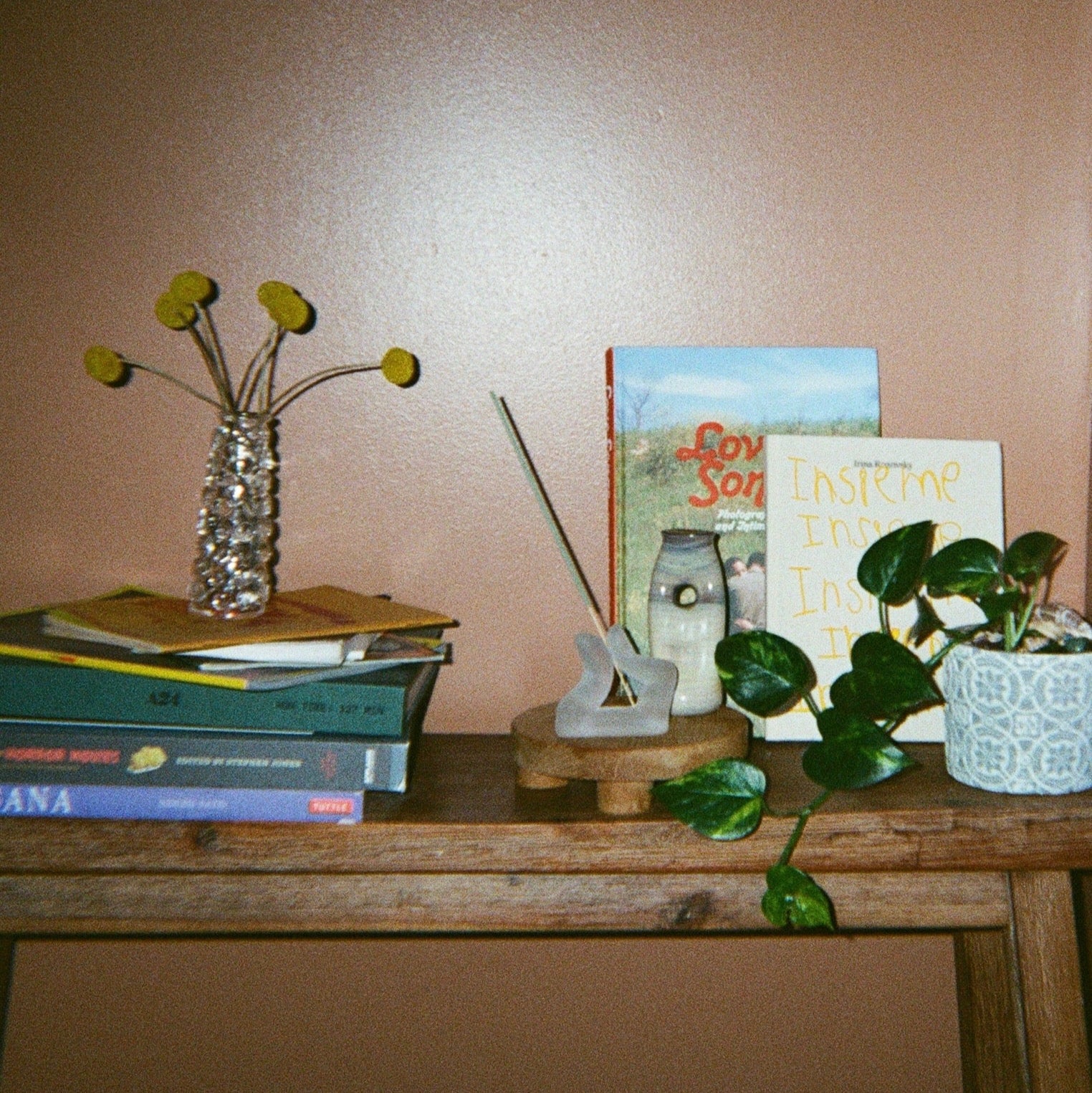 A clear glass flower vessel holding yellow billy buttons, a finger incense holder, another small vessel, a plant, and stacked books on a wooden bench against a brown wall. 