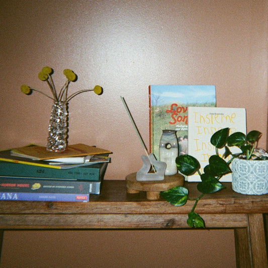 A clear glass flower vessel holding yellow billy buttons, a finger incense holder, another small vessel, a plant, and stacked books on a wooden bench against a brown wall. 