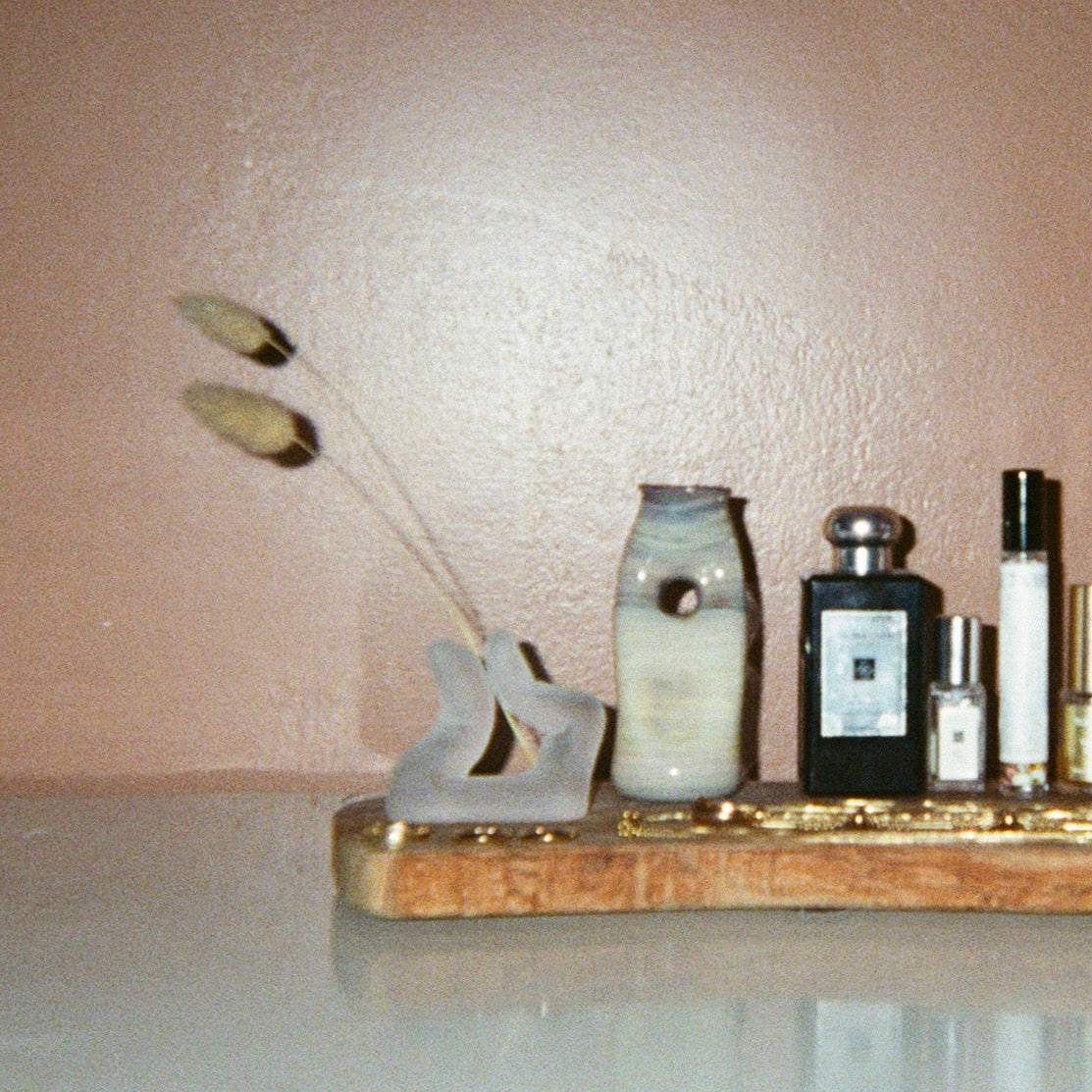 a finger shaped incense holder holding two pieces of dried bunny tail, a free-form vessel with blues, purples, and yellows and perfume bottles on top of a wooden tray. 