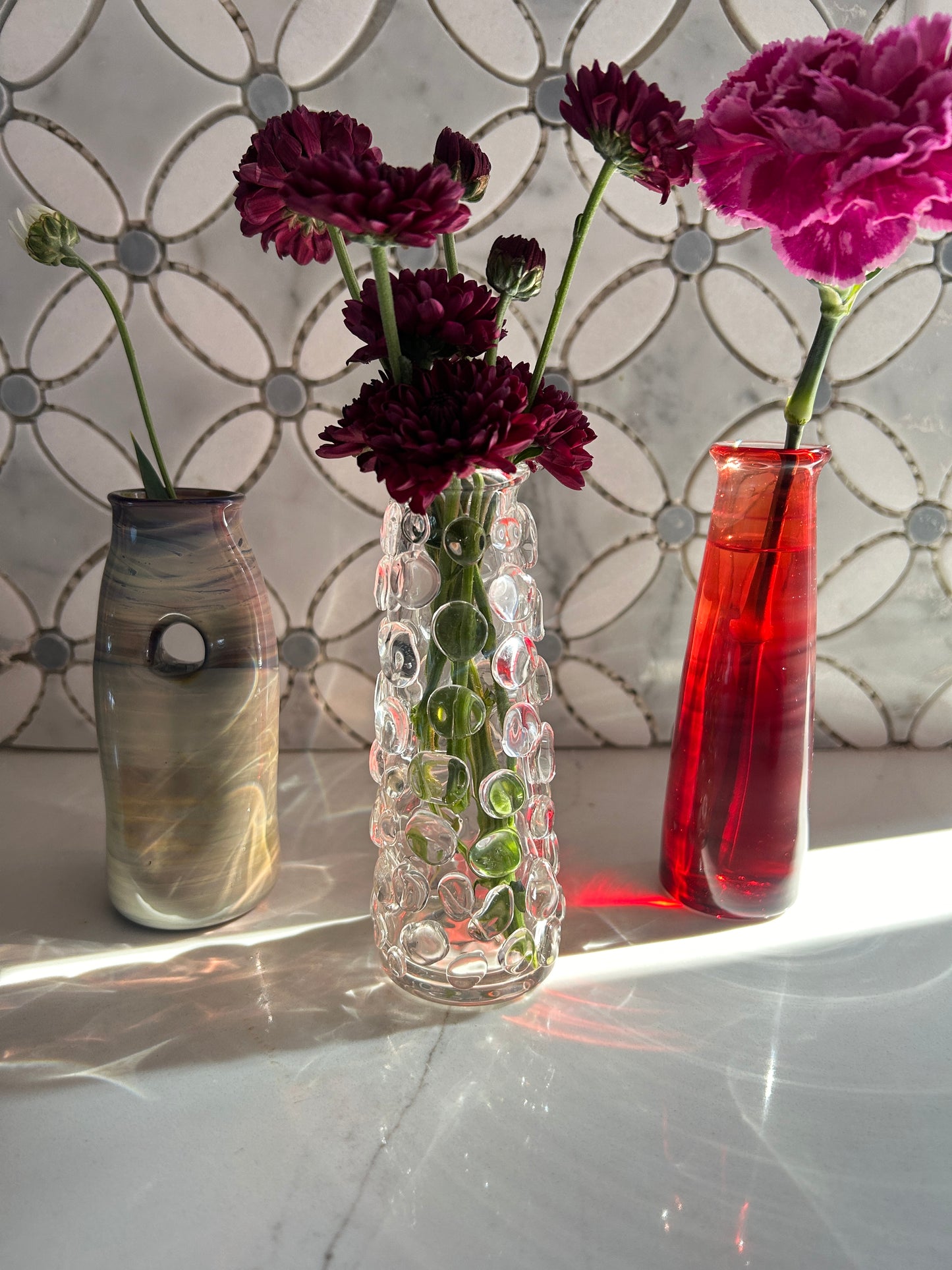 Three flower vessels in varying heights, shapes, and colors holding flowers on a white marble countertop. 