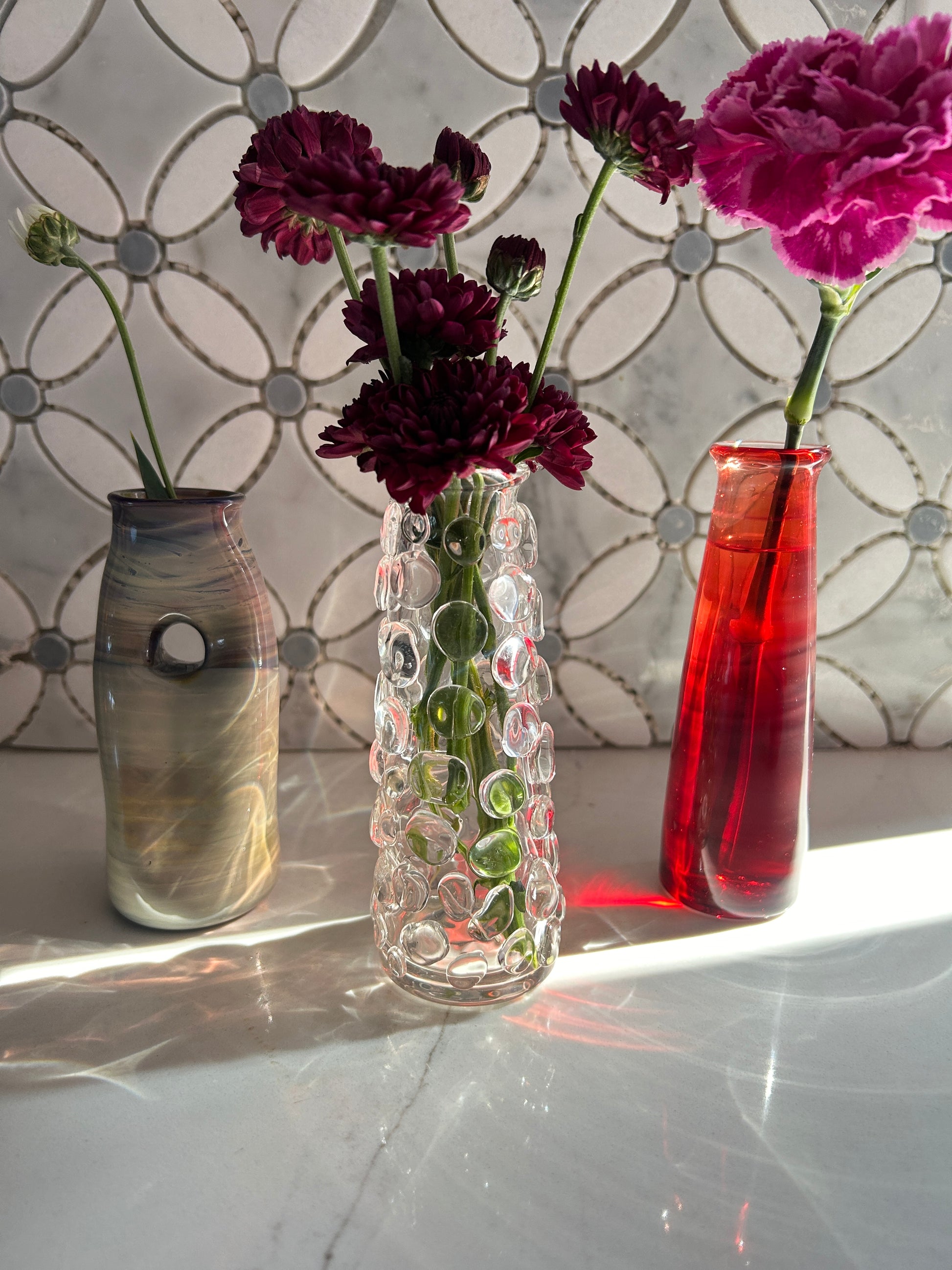Three flower vessels in varying heights, shapes, and colors holding flowers on a white marble countertop. 