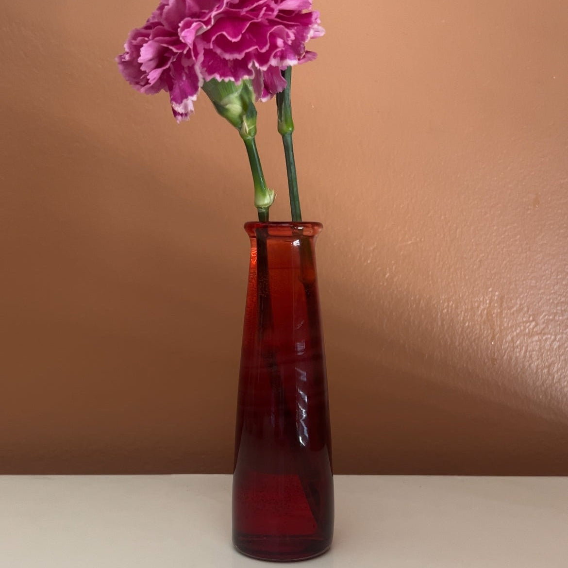 A red flower vessel holding two pink carnations against a brown background. 