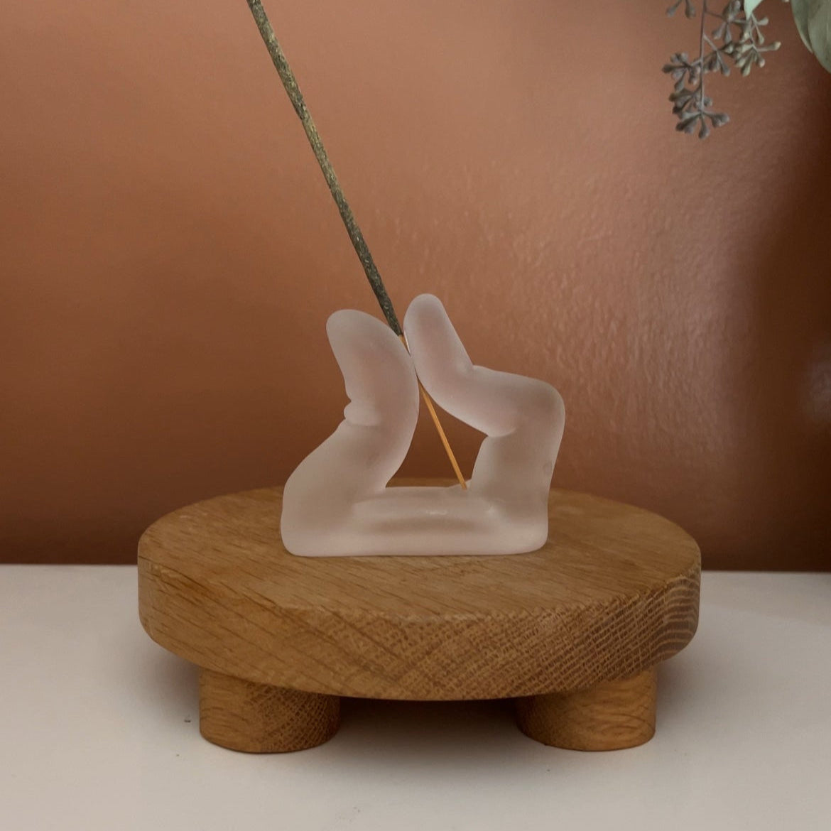 An incense holder shaped like two fingers pinching together on a wooden riser against a brown wall. 