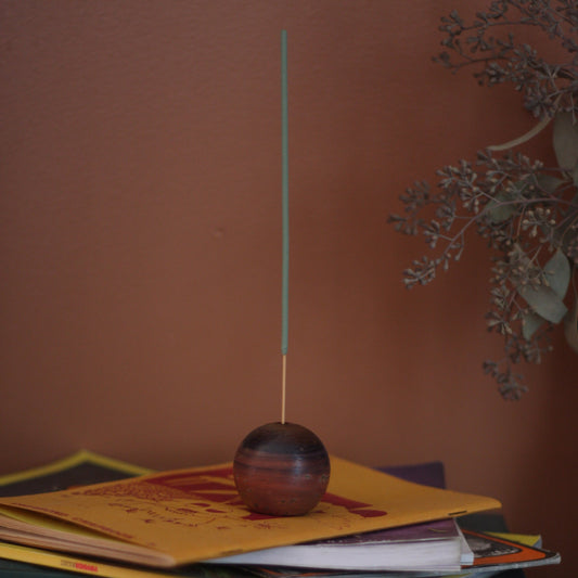 A glass circular incense holder colored with dark purple and terracotta and reddish hues holds a green incense stick. The incense holder sits on a stack of books against a brown wall with dried green leaves in the frame. 