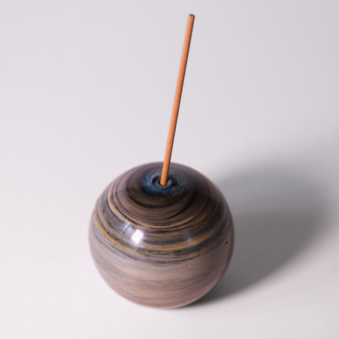 A circular glass incense holder featuring dark blue, greens, yellows and light blue striations. the holder has an incense stick in the center.