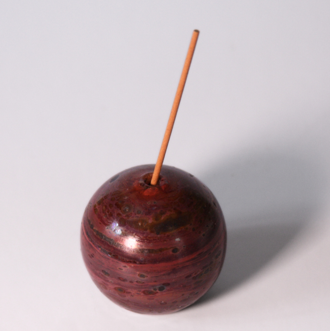 A circular glass incense older with magenta, dark maroon, brown, and pink striations that make the circle look planetary. An incense stick is in the holder. 