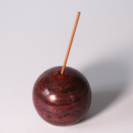 A circular glass incense older with magenta, dark maroon, brown, and pink striations that make the circle look planetary. An incense stick is in the holder. 